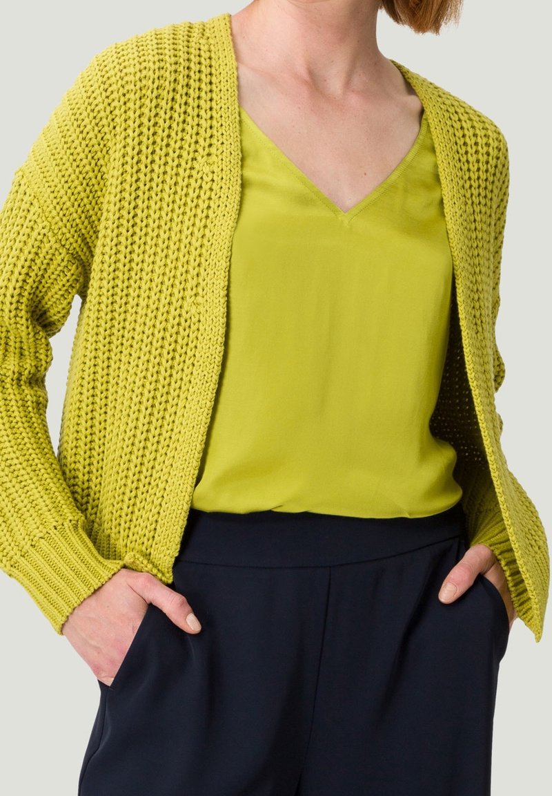 Femme portant un cardigan en maille vert citron par-dessus un haut à col en V assorti, les mains dans les poches d'un pantalon bleu marine, vue du cou jusqu'aux hanches.