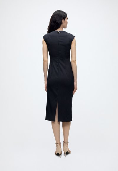 Femme debout, de dos, vêtue d'une robe midi noire ajustée sans manches à fines rayures, avec une fente dans le dos et des sandales beige à talons hauts.