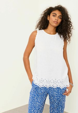 Kvinde med krøllet hår iført en hvid ærmeløs top med hulmønster i kanten og blå bukser med hvidt bølget stribemønster, stående med hænderne i lommerne.