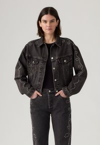Woman wearing black denim jacket and jeans with white studded swirl patterns, hands in pockets, against plain light background.