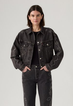 Donna che indossa una giacca di denim nera e jeans con motivi a spirale bianchi borchiati, con le mani nelle tasche, su uno sfondo chiaro e semplice.