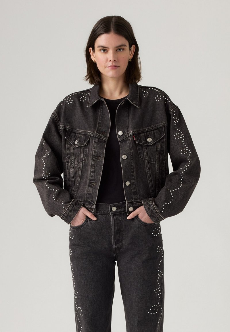 Woman wearing black denim jacket and jeans with white studded swirl patterns, hands in pockets, against plain light background.