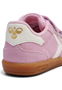 Różowy zamszowy sneaker z białymi akcentami, z złotym logo na pięcie, teksturowaną gumową podeszwą i wyściełanym językiem dla komfortu.