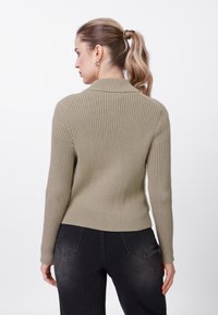 Πλεκτό πουλόβερ σε ελιά πράσινο με ραβδωτή υφή, εφαρμοστή σιλουέτα και ψηλό λαιμό, συνδυασμένο με σκούρο γκρι τζιν.