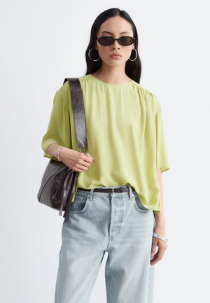 Femme portant un chemisier ample vert citron, un jean bleu clair, une ceinture noire, des lunettes de soleil foncées, des boucles d'oreilles créoles et un sac à épaule marron foncé.