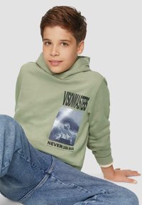 Groene hoodie met een grote grafische print van een persoon in een capuchon en de tekst "Kijk NOOIT terug." Gecombineerd met blauwe jeans.