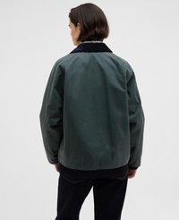 Casaco encerado verde com colarinho de veludo cotelê azul marinho. Apresenta um corte relaxado e textura suave, costuras visíveis e um comprimento intermédio.