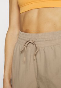 Närbild av en person som bär beige byxor med elastisk midja och knytband samt en orange kort topp som visar magen.