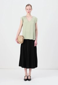 Lys grønn v-hals topp med korte ermer, kombinert med et svart plisserte midi skjørt. Holder en brettet beige clutch, iført svarte leiligheter.