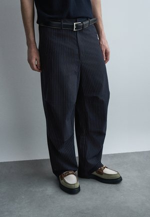 Άνδρας που φοράει σκουρόχρωμο παντελόνι με ψιλή ρίγα, μαύρη ζώνη και πράσινα και λευκά μοκασίνια με καφέ δέρμα και χρυσή αλυσίδα, στέκεται σε γκρι πάτωμα.