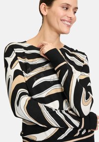 Schwarzer Pullover mit langen Ärmeln, der abstrakte beigen und weißen Wellenmuster zeigt und auf der Vorderseite mit kleinen Nieten verziert ist. Weicher Strickstoff.