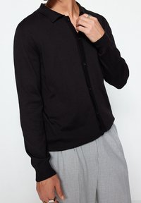 Svart stickad cardigan med en enkel krage, knappstängning och ribbade ärmslut, kombinerad med ljusgrå byxor.