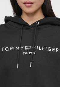 Juodas medvilninis džemperis su kapuce, kuris turi balta "TOMMY HILFIGER" logotipą ir užrašą "EST. 1985". Apima raištelius ir sidabrinius akį akcentus.