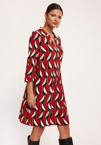 Czerwona sukienka z geometrycznym wzorem w bieli, różu i brązie, z kołnierzykiem i rękawami o długości trzech czwartek. Teksturowany materiał.