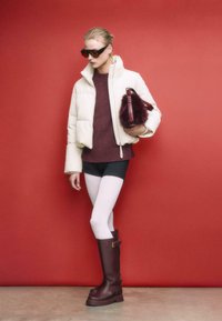 Weiße gepolsterte Jacke, burgunderfarbener Pullover, schwarze Shorts, weiße Strumpfhosen und braune kniehohe Stiefel. Hält eine Felltasche. Roter Hintergrund.