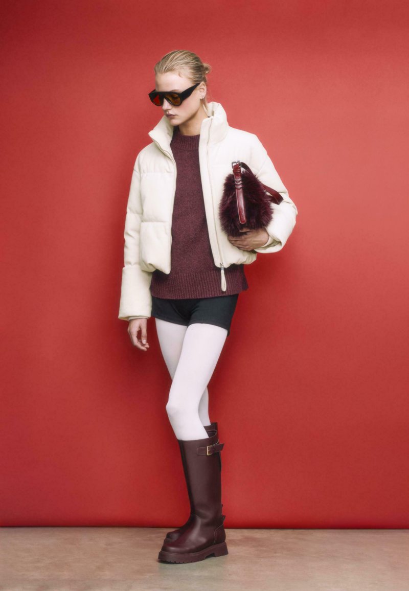 Weiße gepolsterte Jacke, burgunderfarbener Pullover, schwarze Shorts, weiße Strumpfhosen und braune kniehohe Stiefel. Hält eine Felltasche. Roter Hintergrund.