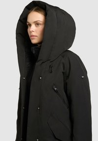 Sort parka med oversized hætte, har en tekstureret overflade, front snap-knapper og lynlåslommer på siden og brystet.