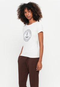 T-shirt de algodão branco com decote redondo e mangas curtas, apresentando um design de logótipo de âncora azul-marinho em padrão circular na parte da frente.