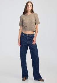 Kobieta stoi w brązowym, krótkim dzianinowym swetrze z detalami z koralików, ciemnych dżinsach typu wide-leg oraz brązowych butach w wzory na jednolitym szarym tle.