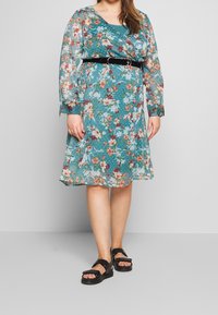 La robe florale présente un fond turquoise, un tissu léger et transparent, une coupe fluide, et une ceinture noire mettant en valeur la taille. Des sandales noires complètent le look.