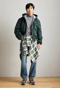 Zelena bomber jakna, sivi turtleneck, karirana košulja vezana oko struka, plave traperice i patike s uzorkom i vezicama; ležeran višeslojan izgled.