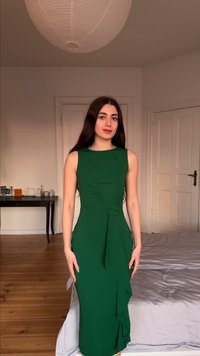 Γυναίκα με μακριά σκούρα μαλλιά, φορώντας εφαρμοστό πράσινο φόρεμα χωρίς μανίκια, στέκεται σε ένα υπνοδωμάτιο με ξύλινο πάτωμα και λευκούς τοίχους.