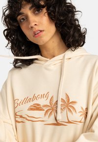 Felpa crema con cappuccio, dotata di cordino regolabile, caratterizzata da grafiche di palme arancioni e dal logo "Billabong". Tessuto morbido e texture piacevole.