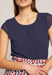 Haut à manches courtes bleu marine avec un col rond, fabriqué en tissu lisse. La main du modèle repose sur une jupe à motifs présentant des formes rouges et marines.