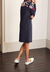 Abito felpa blu marino con vivace ricamo floreale sulle spalle, caratterizzato da una vestibilità rilassata e lunghezza al ginocchio, abbinato a sneakers bianche.