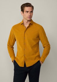 Camisa de botón de manga larga color mostaza con cuello clásico y botones en el frente, hecha de tela texturizada y con un corte ajustado.
