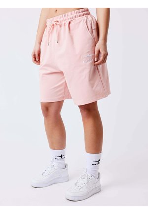 Persoon draagt lichtroze short met trekkoord, witte sokken met zwart logo en witte sneakers, staand op een effen lichte achtergrond.