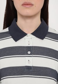 T-shirt polo avec des rayures horizontales bleu marine et blanches, col côtelé, patte de boutonnage avec des boutons de couleur claire et texture en coton douce.