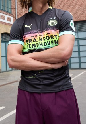 Person mit verschränkten Armen, die ein schwarzes PSV Eindhoven Fußballtrikot mit Brainport Eindhoven Schriftzug und lila Hose trägt, urbaner Hintergrund.