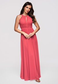 Femme en robe corail rose plissée sans manches avec un décolleté en trou de serrure, debout, les mains jointes, portant des sandales dorées et des boucles d’oreilles.