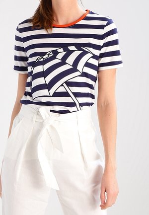 Woman wearing a navy and white striped t-shirt with an umbrella graphic, paired with high-waisted white pants tied at the waist.