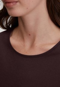 Donkerbruine T-shirt met een brede, ronde halslijn en een gladde textuur, weergegeven tegen de huid. De huid heeft lichte sproeten en een kleine moedervlek.