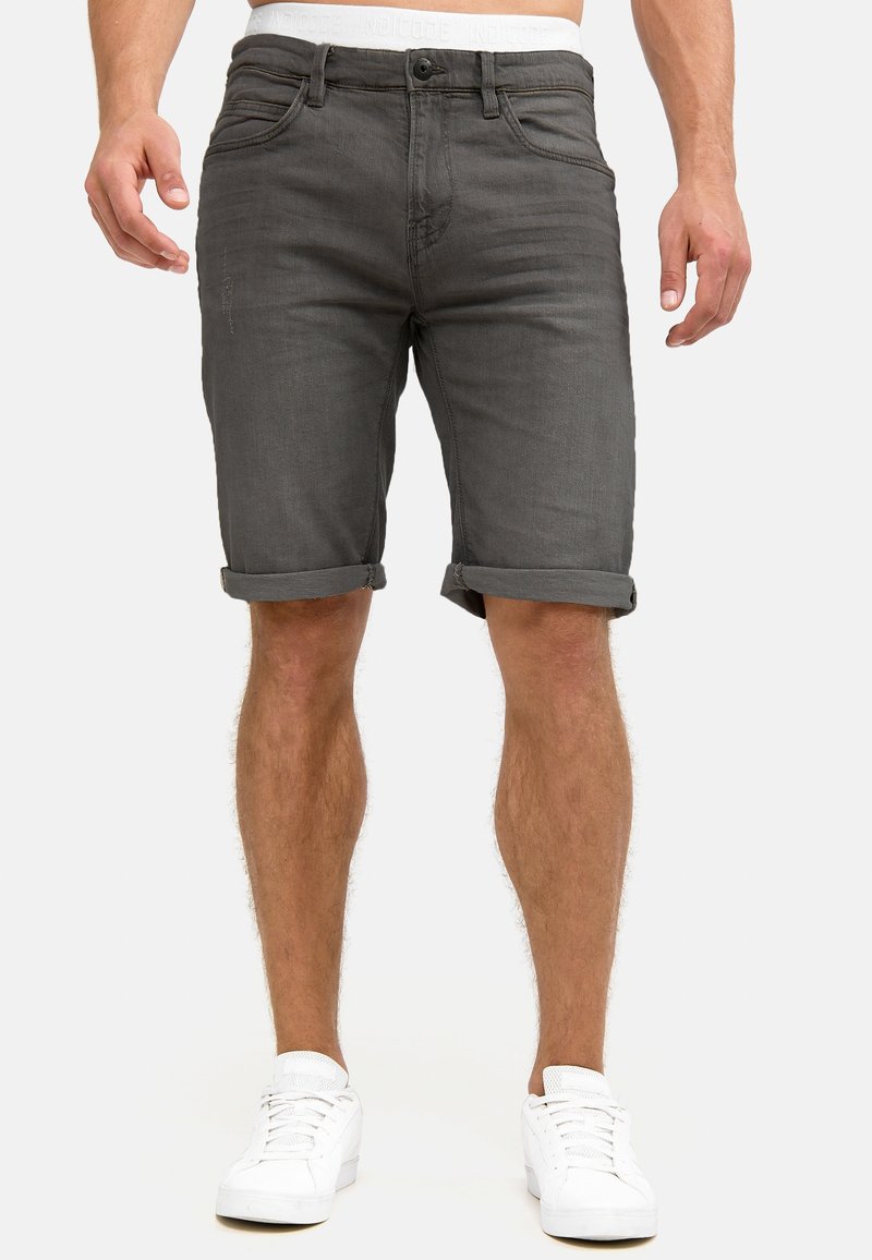 Homme portant un short en jean gris foncé retroussé et des baskets blanches, avec les jambes inférieures visibles, sur un fond uni.