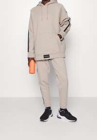 Sweatshirt beige à capuche avec des rayures noires sur les côtés et une poche avant. Associé à un pantalon de jogging fuselé. Chaussures de sport noires, tenant une bouteille d'eau orange.