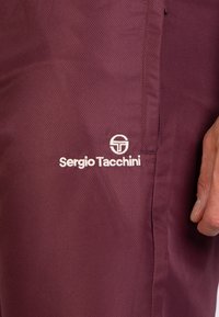 Bordeauxrote Sporthose aus strukturiertem Stoff mit einem weißen "Sergio Tacchini"-Logo an der unteren Beinseite. Mit geradem Schnitt und Seitentaschen.