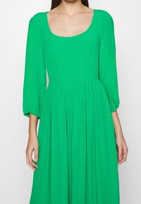 Vihreä mekko, jossa on pyöreä kaula-aukko, pitkät puffihihat ja rypytetty vyötärö. Valmistettu kevyestä kankaasta, jossa on sileä rakenne.