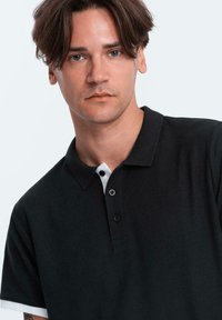 Černé polo tričko s kontrastním bílým límečkem a knoflíkovou légou, vyrobené z texturované tkaniny. Úzce střižený design s krátkými rukávy.