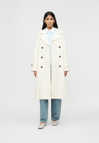 Femme aux longs cheveux bruns portant un trench-coat blanc à double boutonnage, un pull bleu clair, une chemise à col, un jean ample et des chaussures plates blanches.