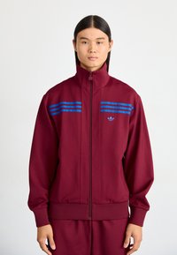 Giacca a track con zip di colore bordeaux, con strisce blu sul petto, polsini e orlo a coste. Presenta un piccolo logo nella parte superiore sinistra.