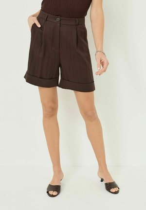 Femme portant un short marron foncé retroussé, un haut sans manches et des sandales à talons ouvertes assorties, debout contre un fond clair uni.