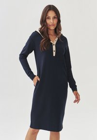 Marineblauwe jurk met lange mouwen en een capuchon, voorzien van een beige knoopsluiting en zakken. Glad, rekbaar materiaal met een ontspannen pasvorm.