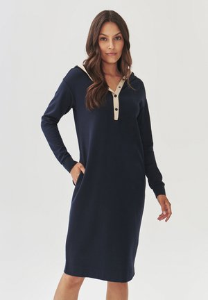 Abito blu navy con maniche lunghe e cappuccio, caratterizzato da una placca di bottoni beige e tasche. Tessuto liscio e elastico con una vestibilità comoda.