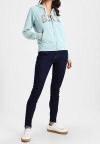 Sweat à capuche bleu clair zippé avec un logo gris, associé à un jean skinny foncé et des baskets blanches avec une semelle beige. Texture de tissu lisse.