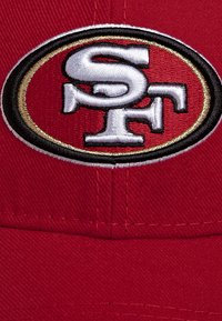 Rote Baseballmütze mit weiß gestickten SF-Initialen innerhalb eines schwarzen und goldenen ovalen Rahmens auf der Vorderseite.