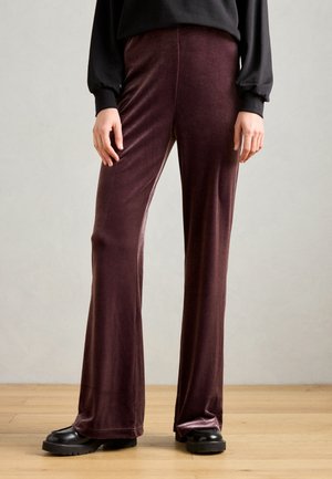 Pantalons évasés en velours violet avec une texture lisse, une taille haute et un léger éclat, associés à des chaussures noires à semelles épaisses.