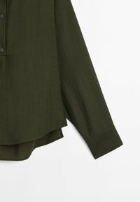 Blouse vert foncé à manches longues avec poignets à boutons et ourlet incurvé, en tissu léger texturé.